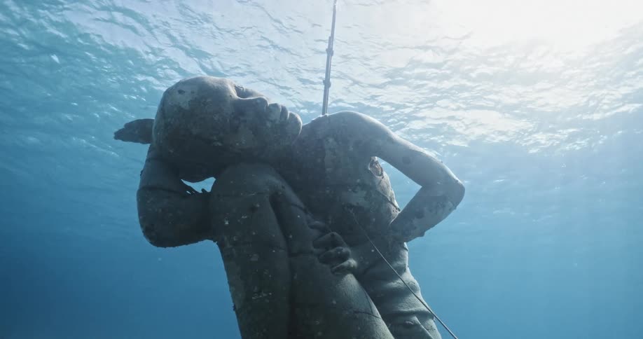 Close up of a statue submerged underwater with fish swimming around in the clear blue ocean water