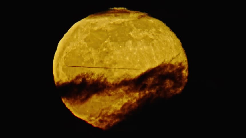 Unreal shot of a commercial jetliner passing in front of a full moon with clouds passing in front of the moon. Shot from 10,000 feet from a helicopter at night