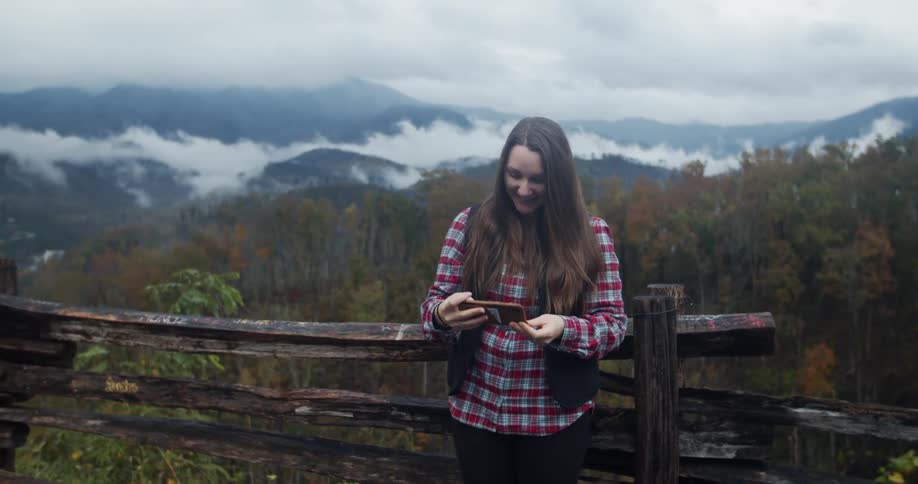 Woman smiles as she stands near worn wooden fence taking selfie with mountains and forest in background
