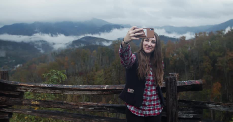 Smiling woman combs fingers through hair as she takes selfie near worn wooden fence with misty forest and mountains in the background