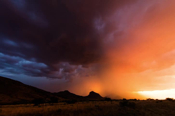 Gray and orange storm clouds shine with lightning strikes over mountains near pasture
