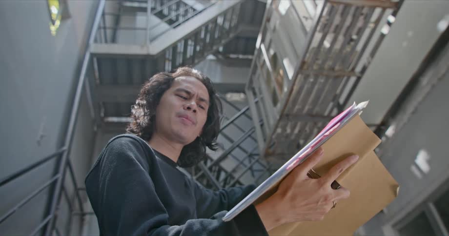 A young man reads from a manila folder full of papers while standing in the stairwell of a building