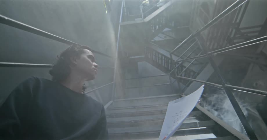 Man stands holding pages of white paper near staircase in fog shrouded area