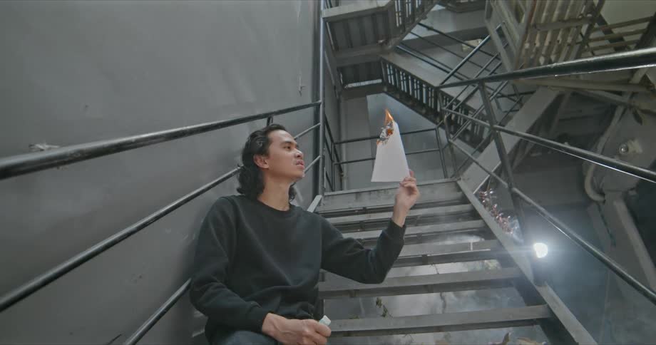 A man sitting in a staircase holding a paper on fire with smoke and ash