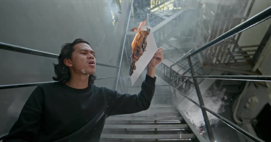 A man sitting in a staircase holding a paper on fire with smoke and ash