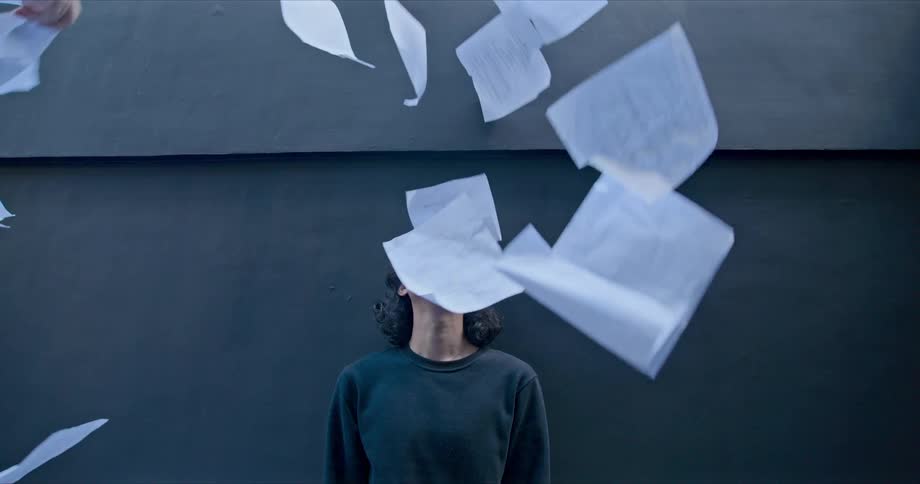 A teenager stands in front of a building and looks up as sheets of paper with handwritten notes fall down from above