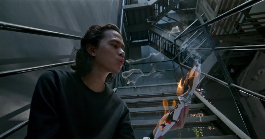 A teenage boy sits in a staircase and blows on papers as they burn