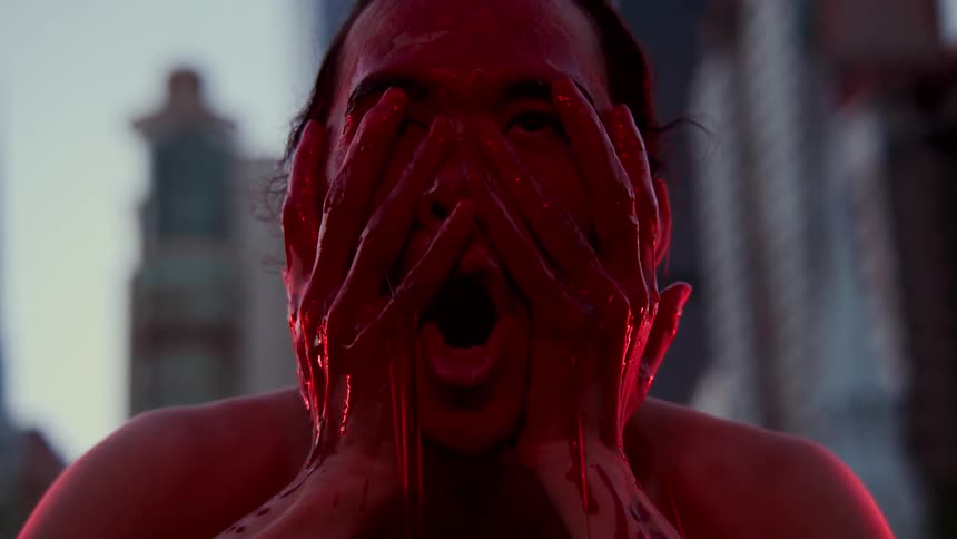 A man putting red paint on his face with buildings the sky and clouds in the background