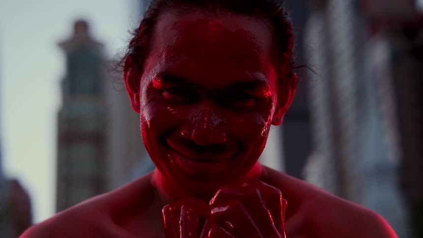 A man smiling with red paint on his face with buildings the sky and clouds in the background