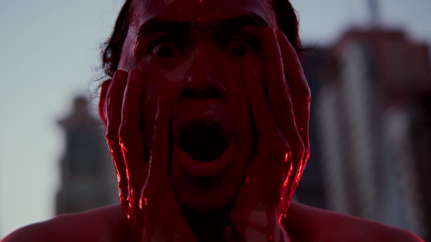 Man stares open mouth as he spreads crimson paint across his face with skyscrapers in background