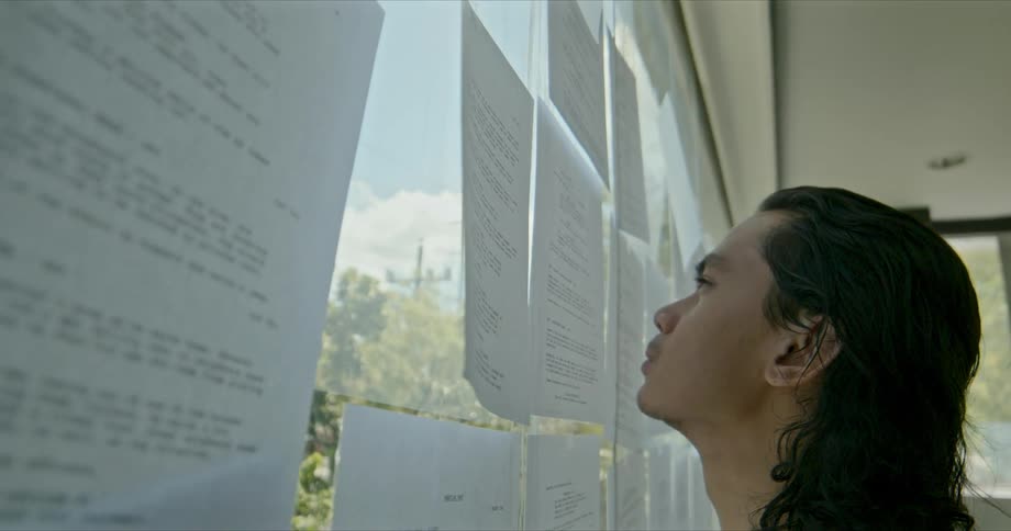 Young man with long hair reading a list on a paper taped on a window