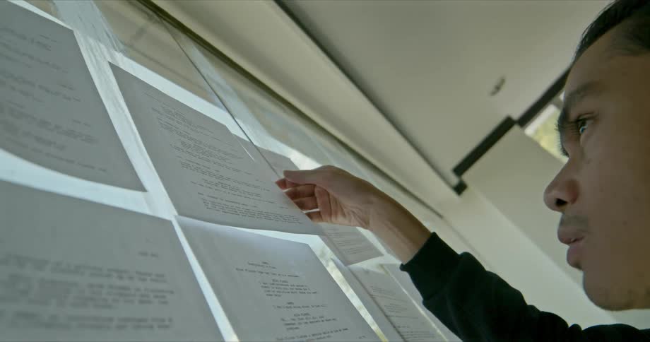A man holds a page of a script and studies it while standing in front of a window covered in script pages