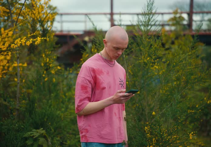 Man with shaved head stands among plants scrolling on mobile phone with bridge in background