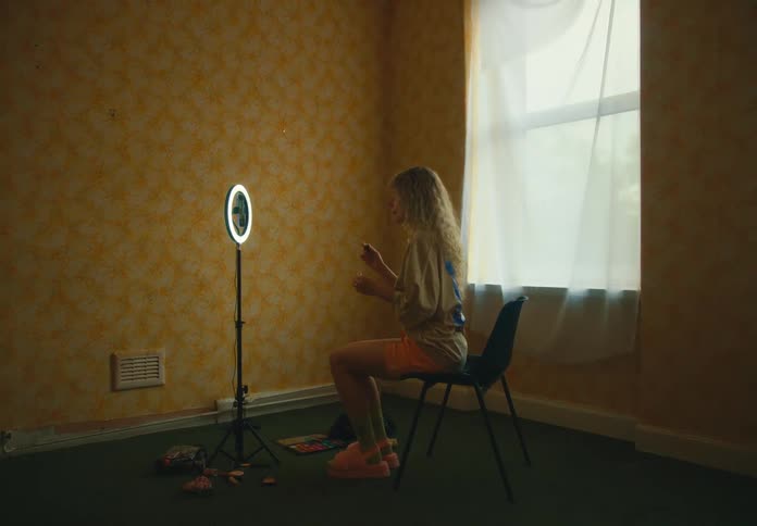 A teenager applies lipgloss in her bedroom while sitting in front of a round mirror on a pole