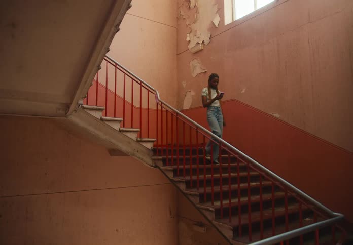 A young woman holding her phone slowly descends a stairwell in an aging building