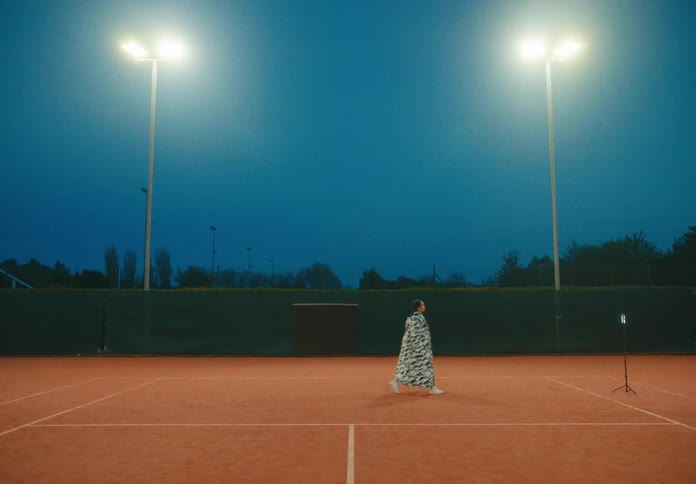 A woman dancing on a tennis court in front of a smartphone on a tripod with trees the sky and clouds in the background