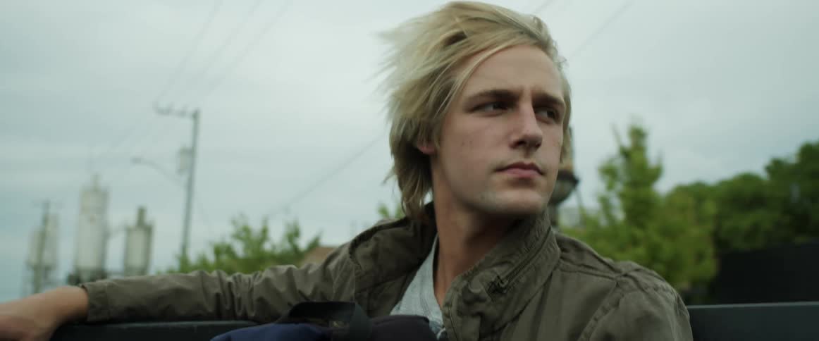 Blonde man sits in back of truck bed driving down street past water tower and transformer