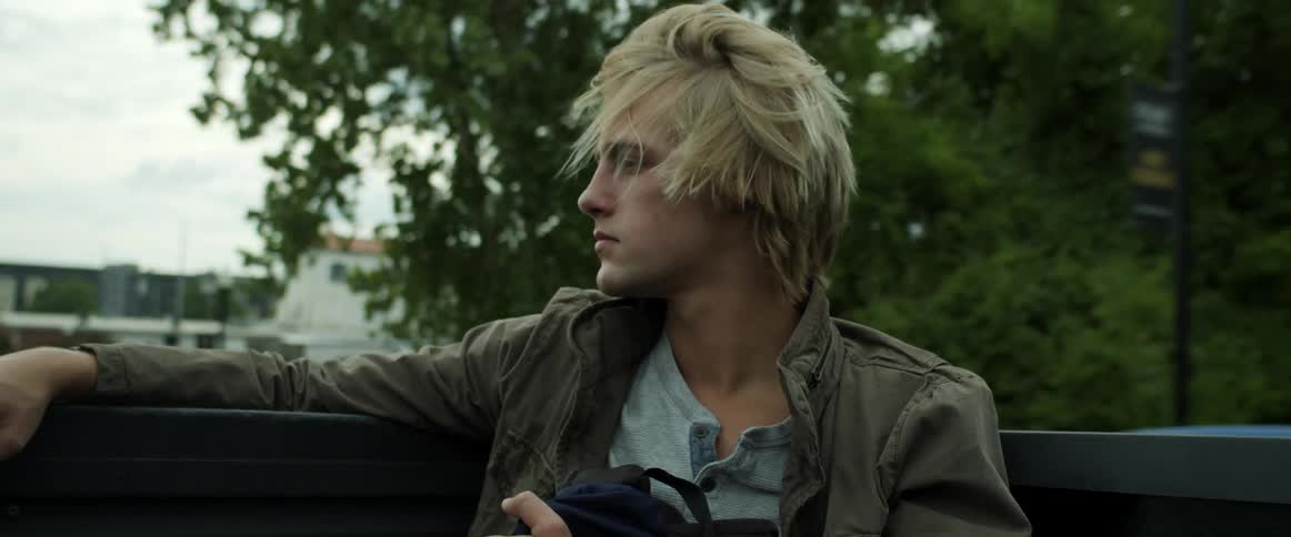 Blonde man sits in truck bed as vehicle drives down road in town