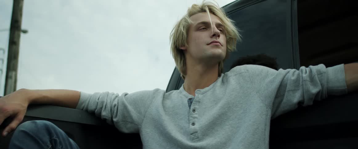 A young man with blonde hair sits in the back of a pickup truck as it is driving along a street