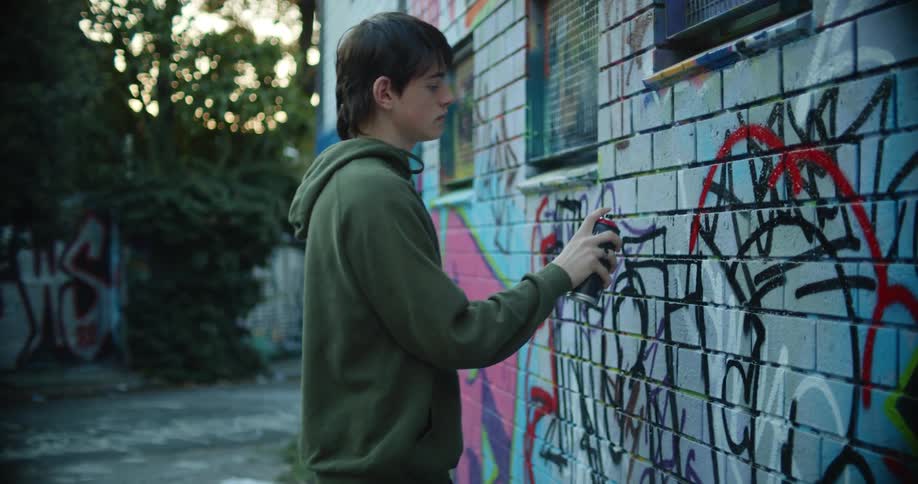 A teenage boy spray paints graffiti on the side of a building