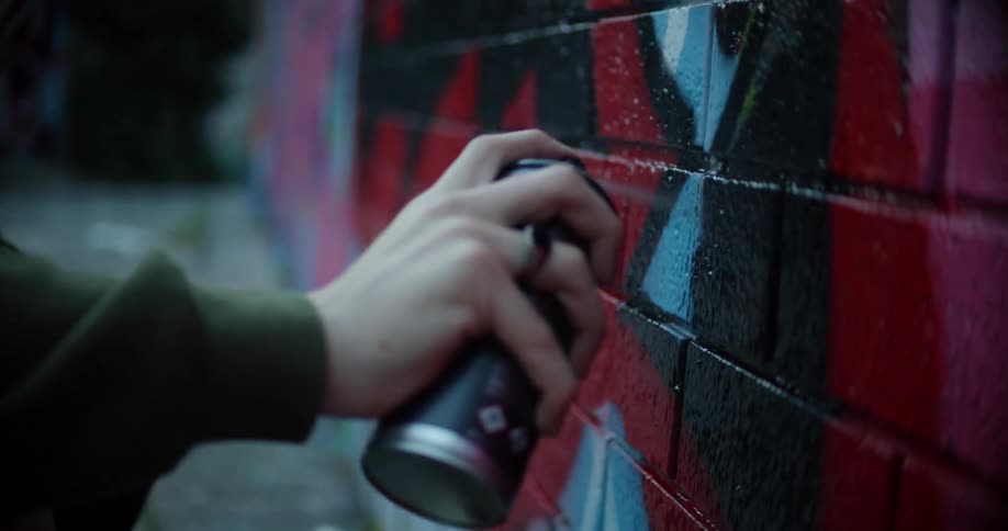 Young graffiti artist wearing hoodie sprays paint onto art on brick wall