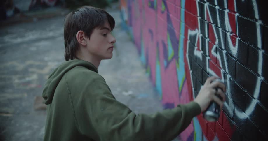 A teenager uses a sketch on paper to spray paint a mural on a brick wall