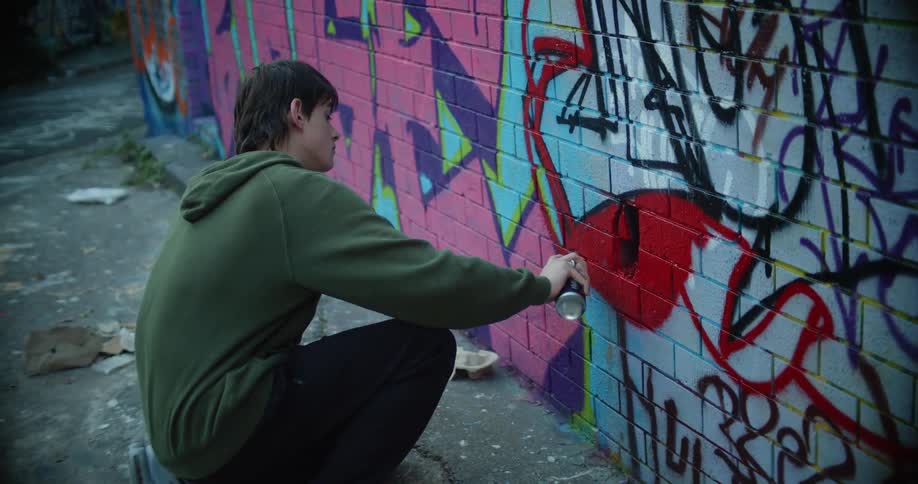 Youth wearing hoodie fills in drawing on graffiti covered wall