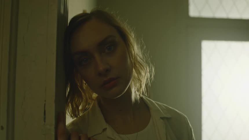 A woman standing by a doorway and staring with a wall and window in the background