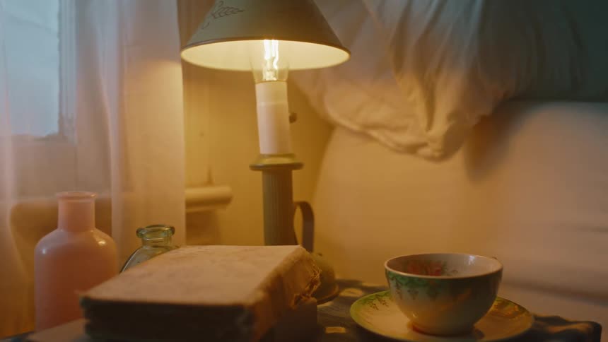 A woman sits on the edge of her bed next to a nightstand with books and a teacup