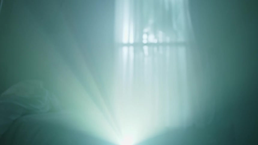 Young woman wearing a white gown walks in front of a projector to sit in a bed on a dimly lit room