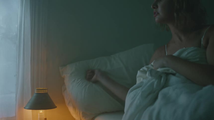 A woman in a white slip climbs into bed and turns off a lamp on the nightstand