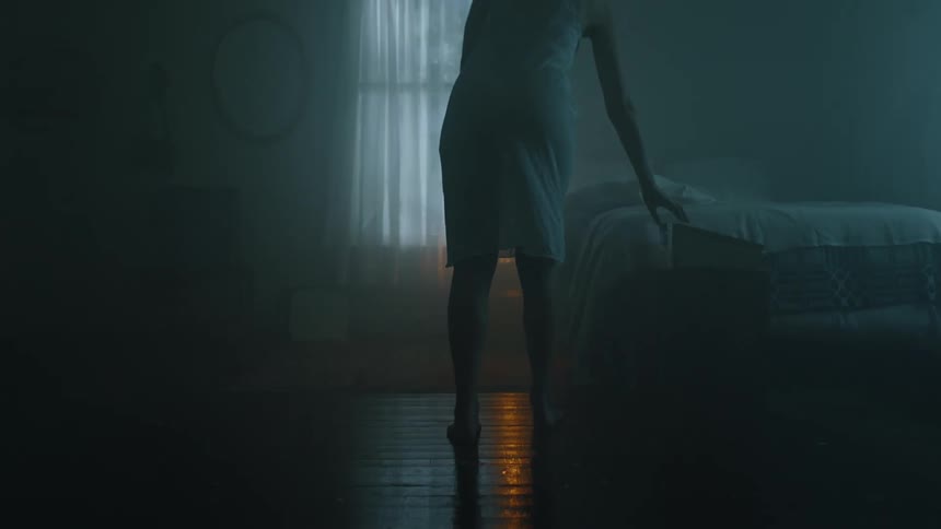 A woman in a night gown walks slowly into a dimly lit bedroom where she turns on a projector before sitting on a bed