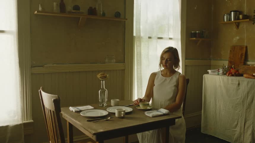 A woman strokes the rim of a tea cup with a finger while sitting lost in thought at a dining table in a house during the day