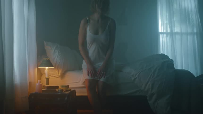 Blonde woman wearing nightgown gets up from bed and walks out of dimly lit room