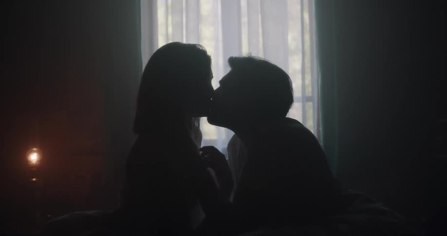 Couple silhouetted by window in room dark dimly lit by lamp recline in bed and kiss