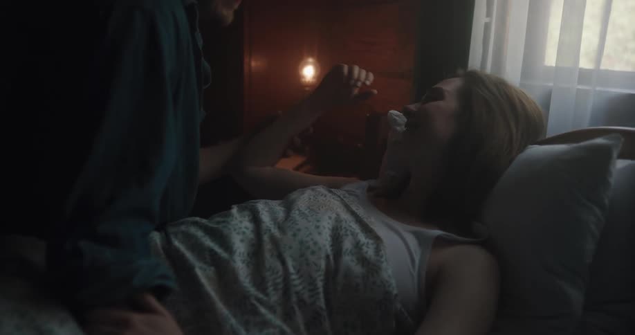 A man and a woman in a bedroom with the woman laying on the bed and the man sitting by her with a wall and window in the background