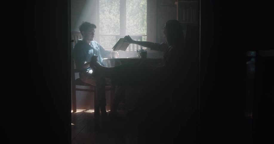 A man and woman sit at a table at home and the man drinks coffee as the woman shows him a book