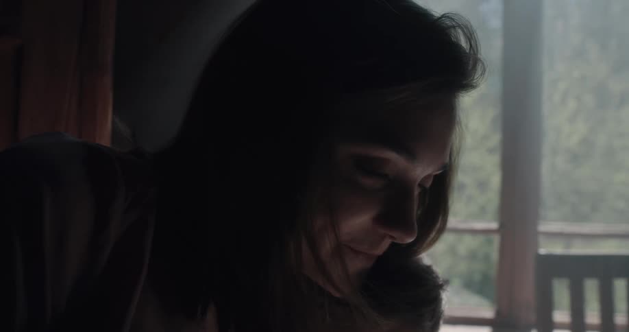 A man and a woman in a room looking at a book with the woman leaning on the man as he sits on a chair with a wall and window in the background
