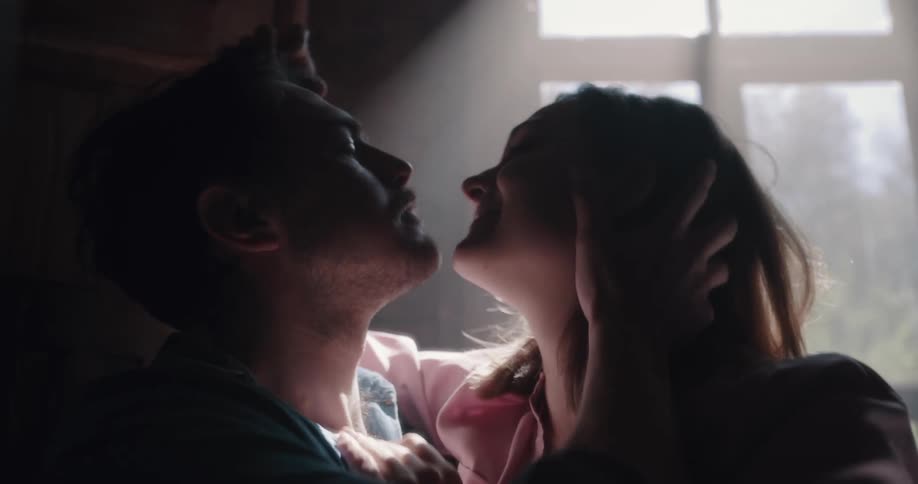 Couple kissing passionately near a window in a dimly lit room on a cabin
