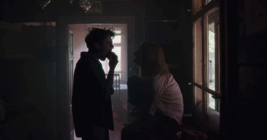 A woman sitting in a corridor tosses an apple to a man who takes a bite before holding it up for her to take a bite as well