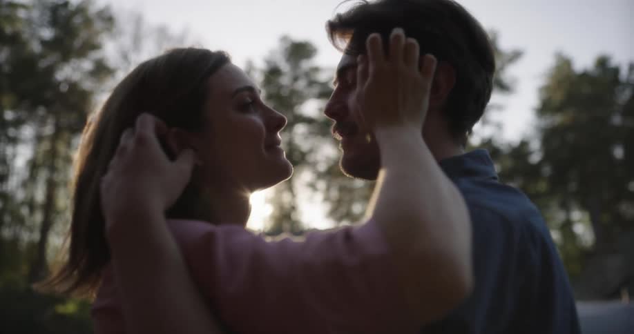 A man and a woman hugging and kissing with trees the sky and clouds in the background