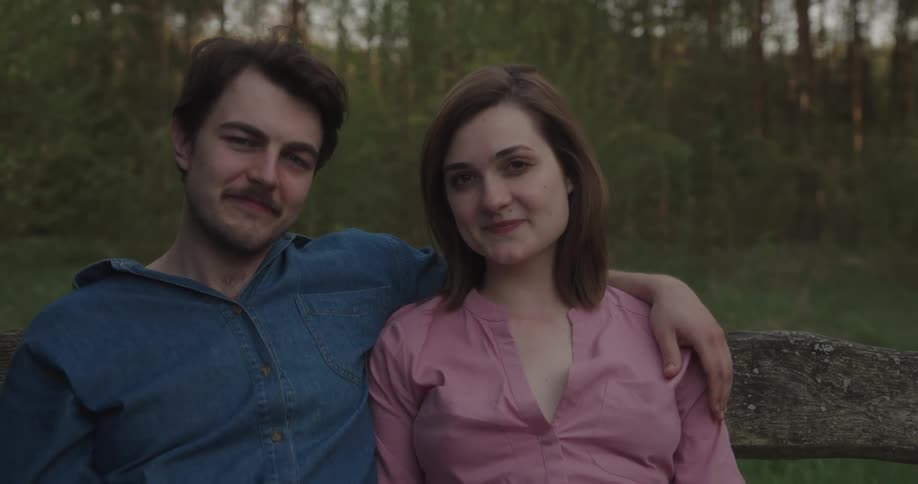 A man and a woman sitting with trees the sky and clouds in the background