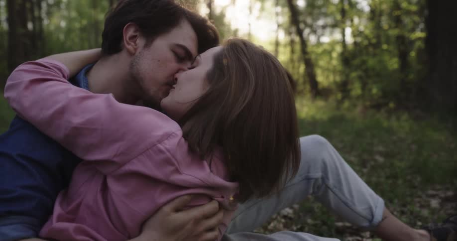 A man and a woman sitting in a forest kissing with trees the sky and clouds in the background