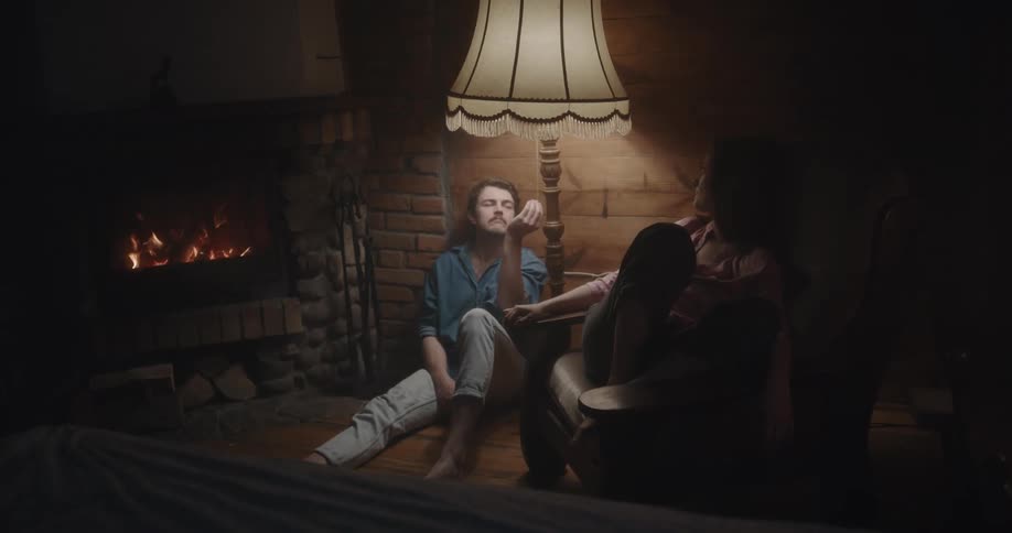 Woman sits on chair next to man sitting between lamp and fireplace as they playfully turn lamp on and off