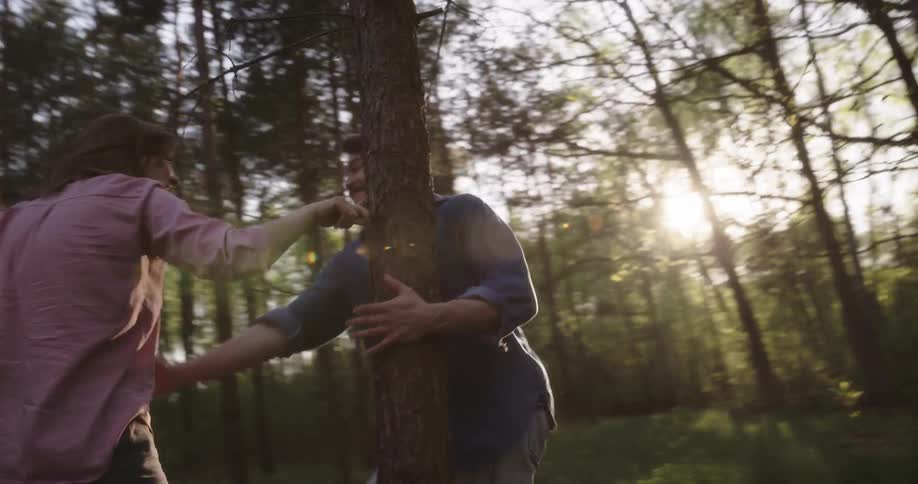 A man chases his girlfriend around a tree in the forest and pulls her in for a kiss