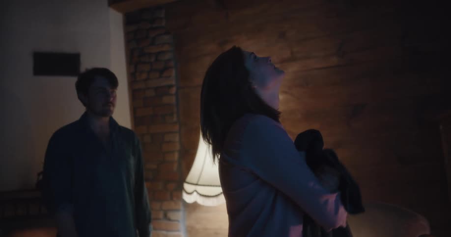A man and a woman in a room with the woman pushing the man with a wall a lamp and fireplace in the background