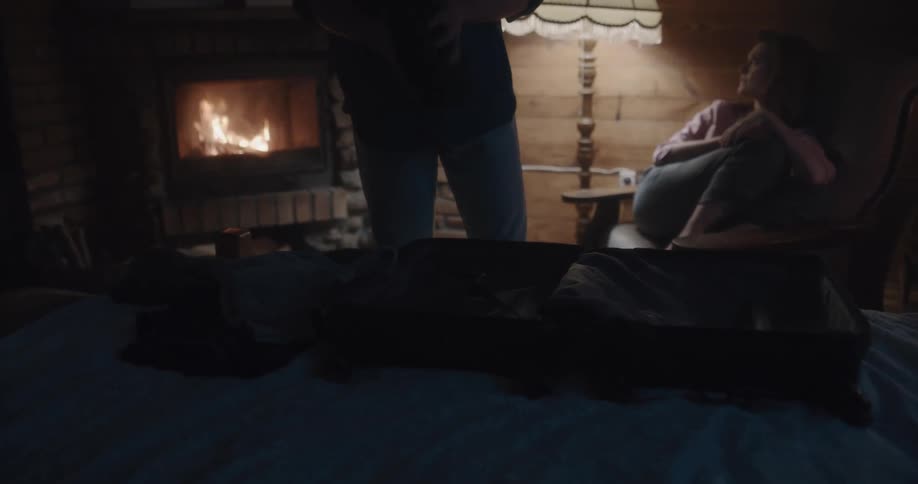 A man unpacks his suitcase on a bed while showing his girlfriend a shirt as she sits in front of a fire