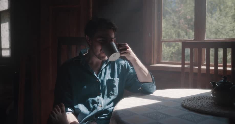 A man in a room sitting on a chair at a table and drinking from a mug