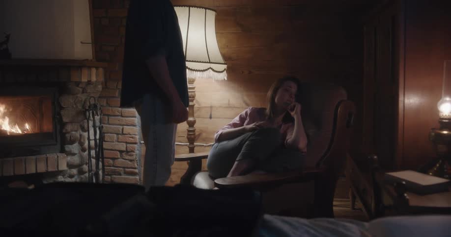 A man and woman are indoors as the woman reclines in a chair