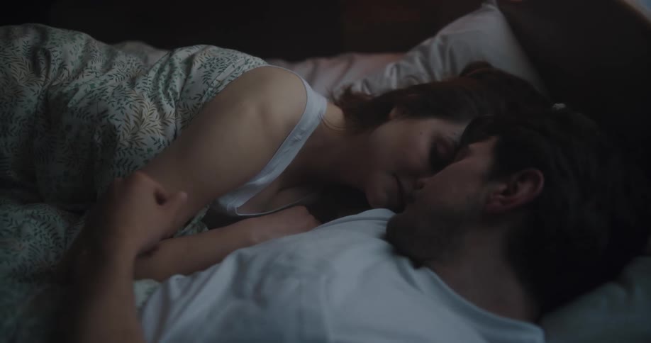 A man and a woman in a bedroom laying on a bed with a wall and a window in the background